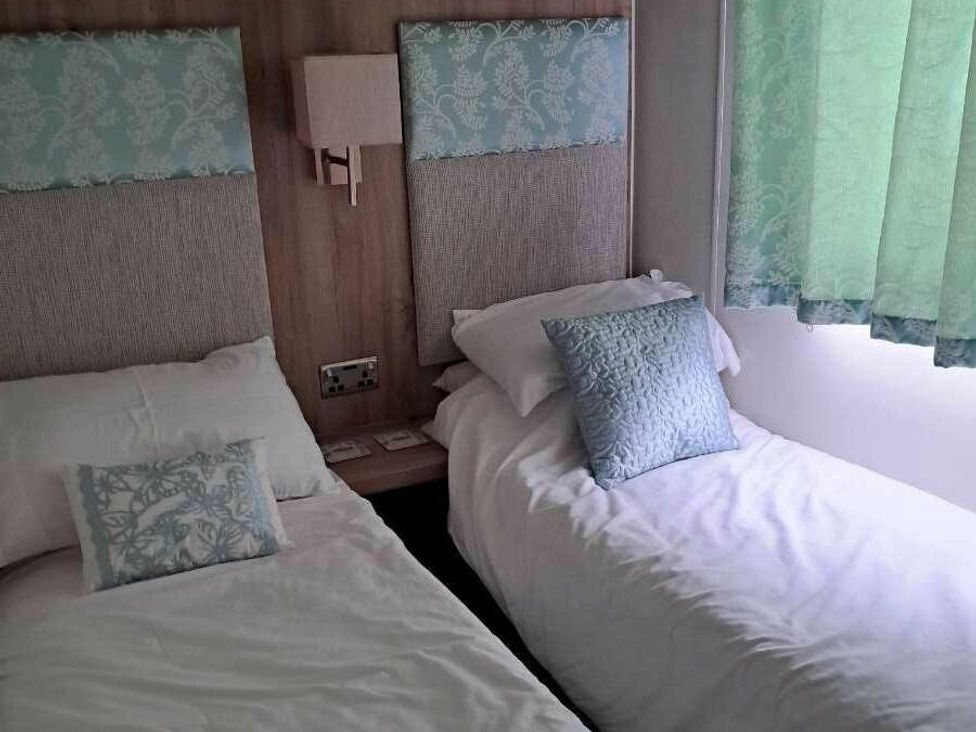 a bedroom with two beds, wall-mounted lamps, and curtains at Golden Sands Holiday Park - Holiday Accommodation 18951, Dawlish