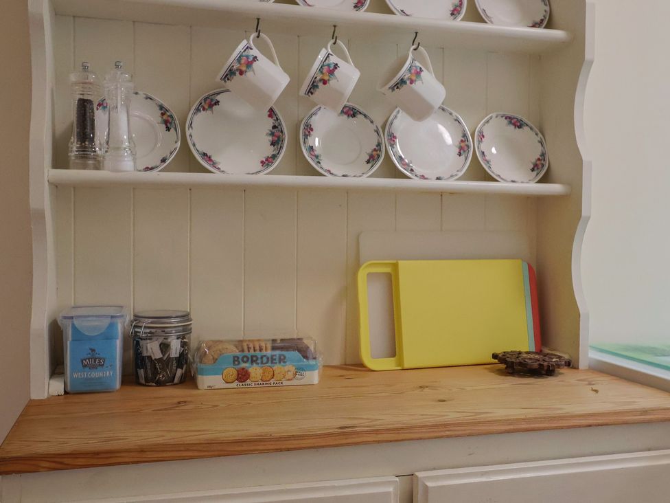 A kitchen shelf with mugs, plates, and containers at Vine Cottage in Burton Bradstock