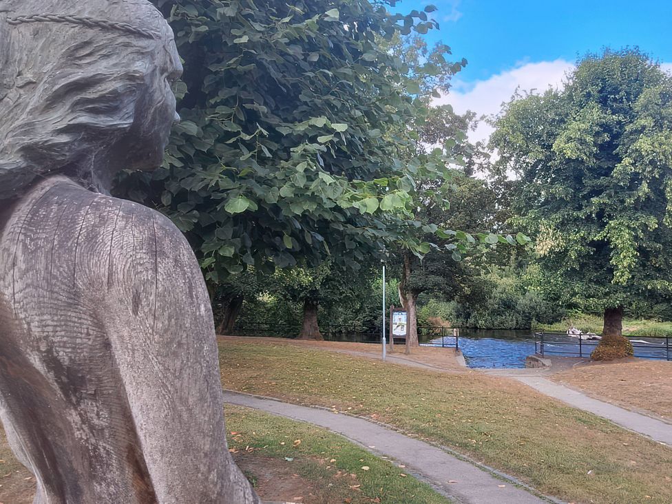 A statue overlooking a river with trees and a pathway at Capel Llani in Llanidloes