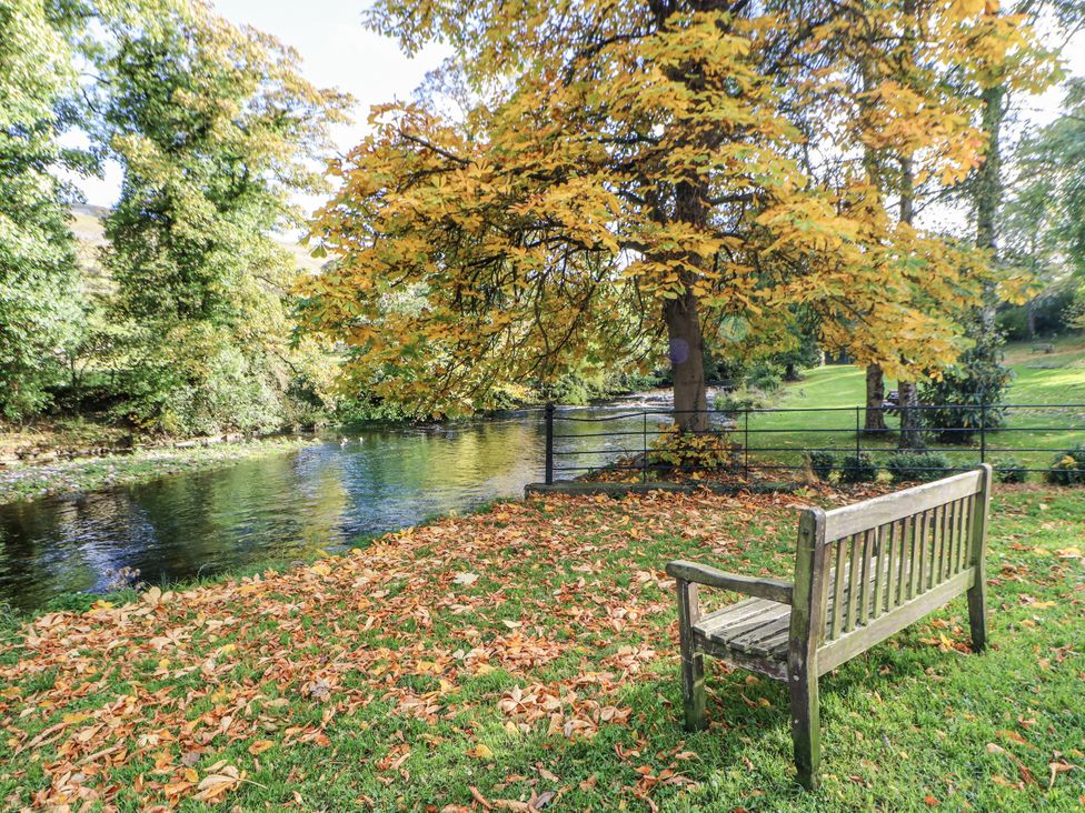 A bench by a river with trees and fallen leaves at Ivy Cottage in West Burton