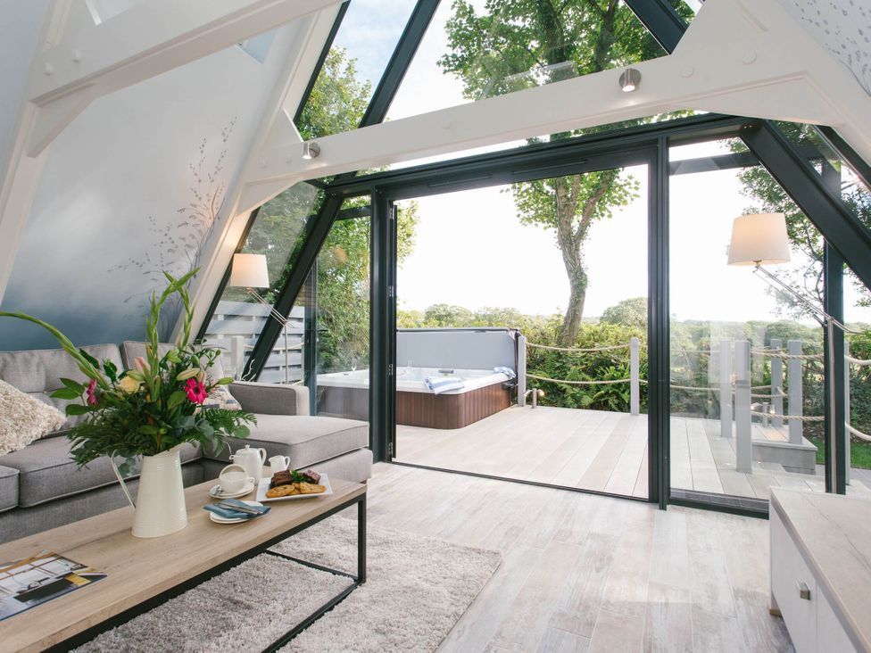 A living room with a sofa and coffee table leading to a deck with a jacuzzi at Glass Lodge in Praze-An-Beeble