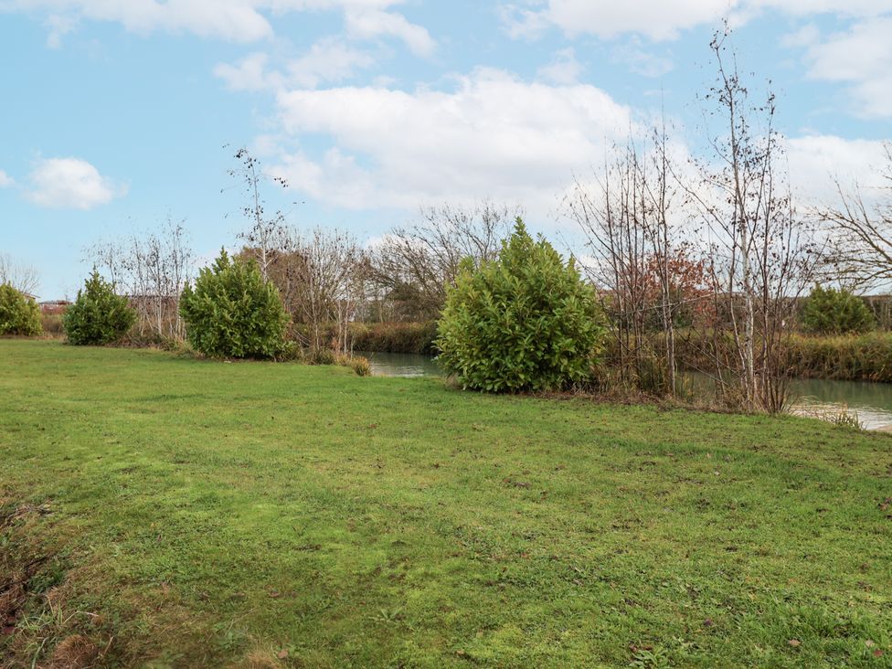 A grassy area near water with shrubs and trees at Lakeside Lodge in Stonham Aspal