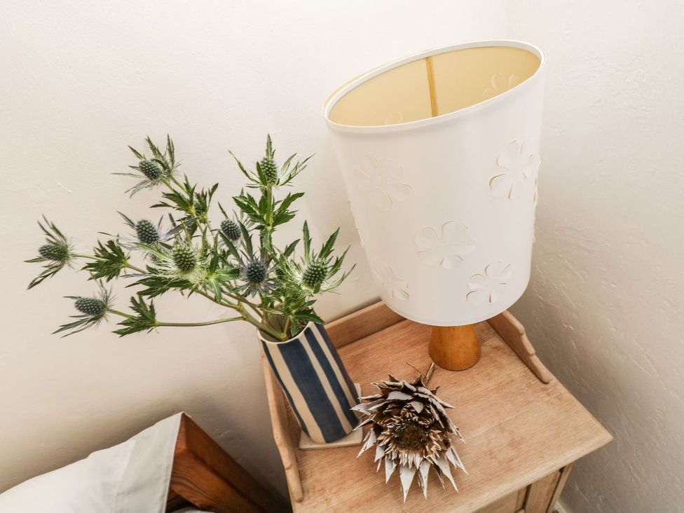A bedside table with a lamp, vase with flowers and a decorative item at Driftwood Cottage in Helston