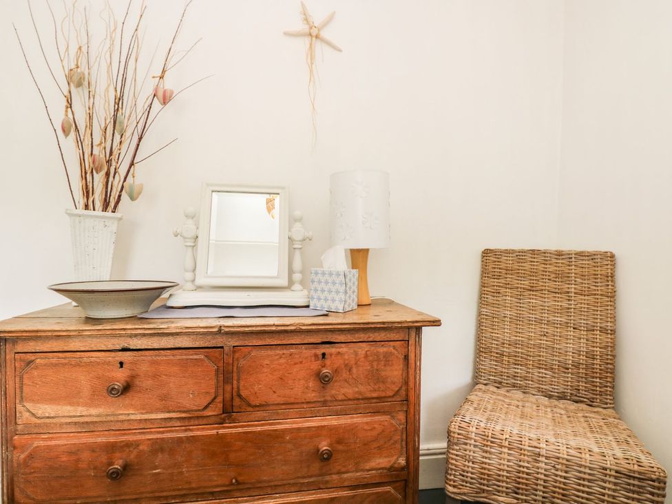 A wooden dresser with a mirror and a wicker chair at Driftwood Cottage in Helston