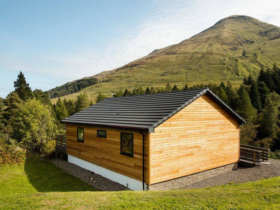 A wooden building with a deck in a mountainous area at Pinemarten Lodge Crianlarich