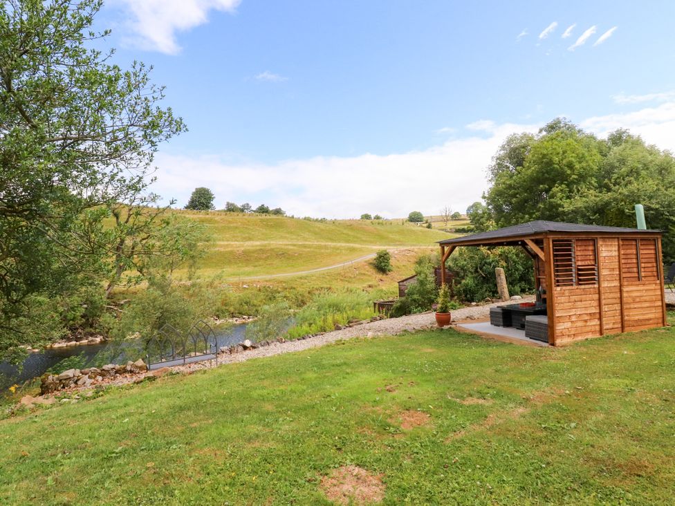 A wooden hut by a river with grass and bushes at Kirk Yett in Linton Falls near Skipton