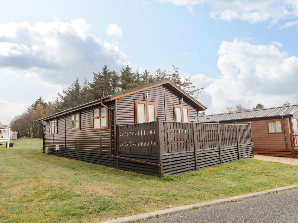 A log cabin with a deck surrounded by grass and trees at Nevaeh Lodge in Longframlington