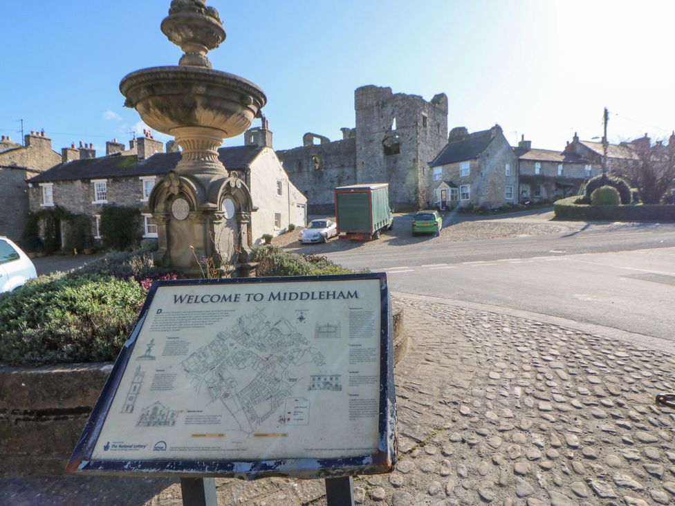 A fountain and sign in a square with buildings and castle ruins in Middleham