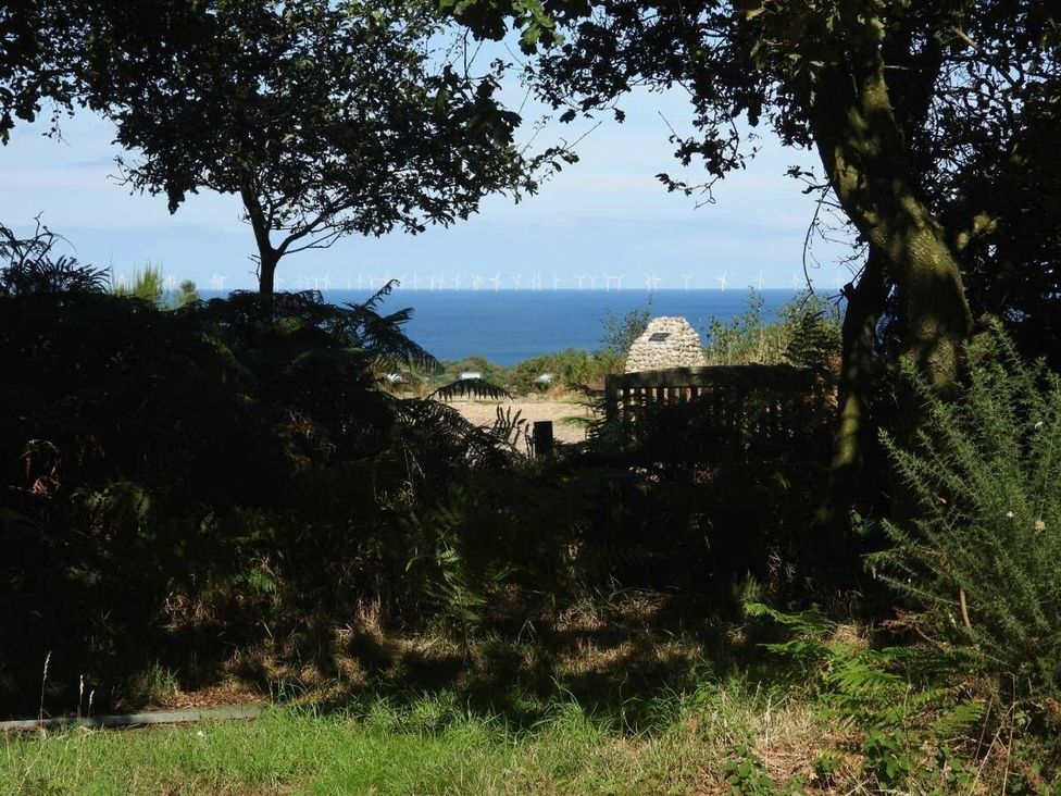A view of the sea with wind turbines from behind trees at Lifes A Beach in Holt