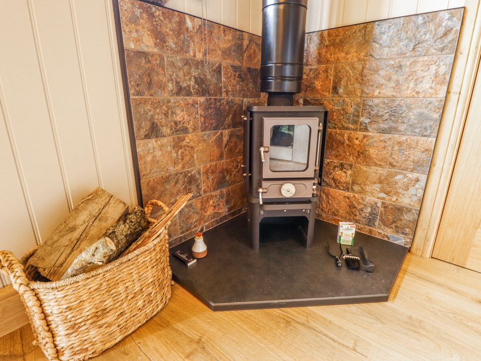 A wood stove with a basket of wood and fireplace tools in a living room at Twit Twoo Shepherds Hut North Somercotes