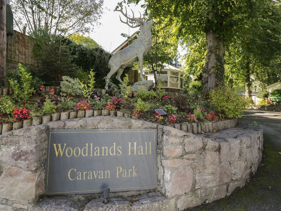 A sign for Woodlands Hall Caravan Park with a deer statue and flowers in Ruthin