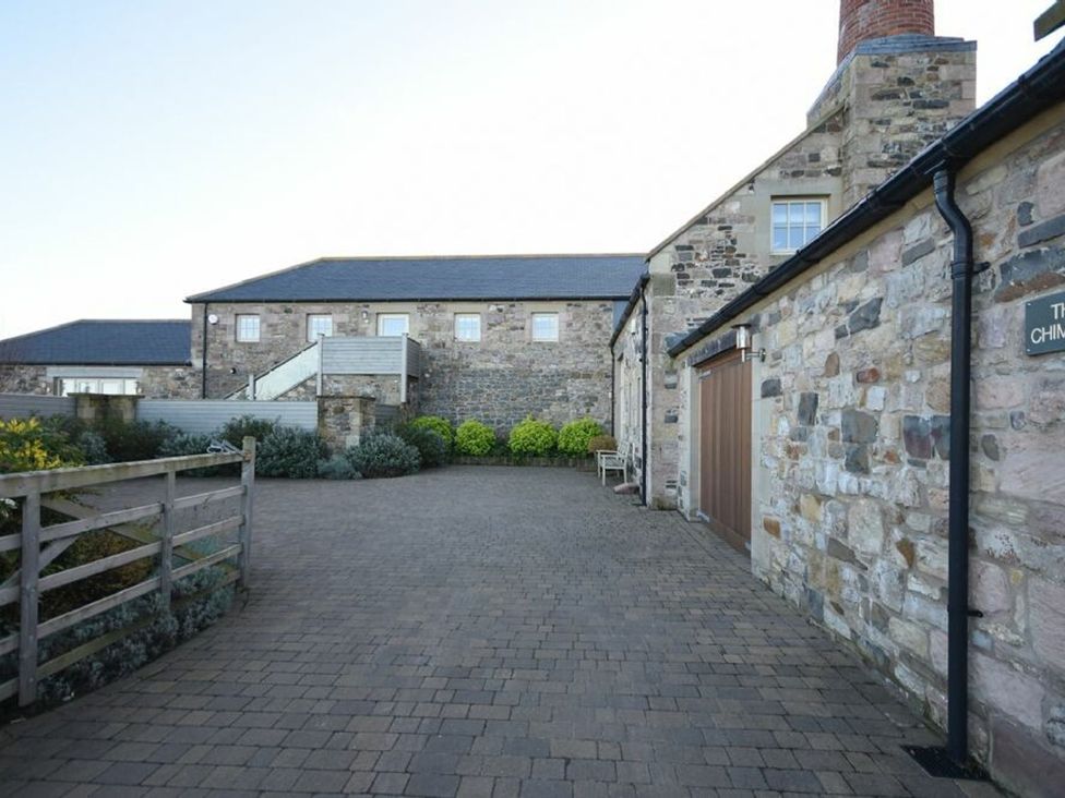 A stone building with a paved driveway and garden area at The Chimney in Bamburgh