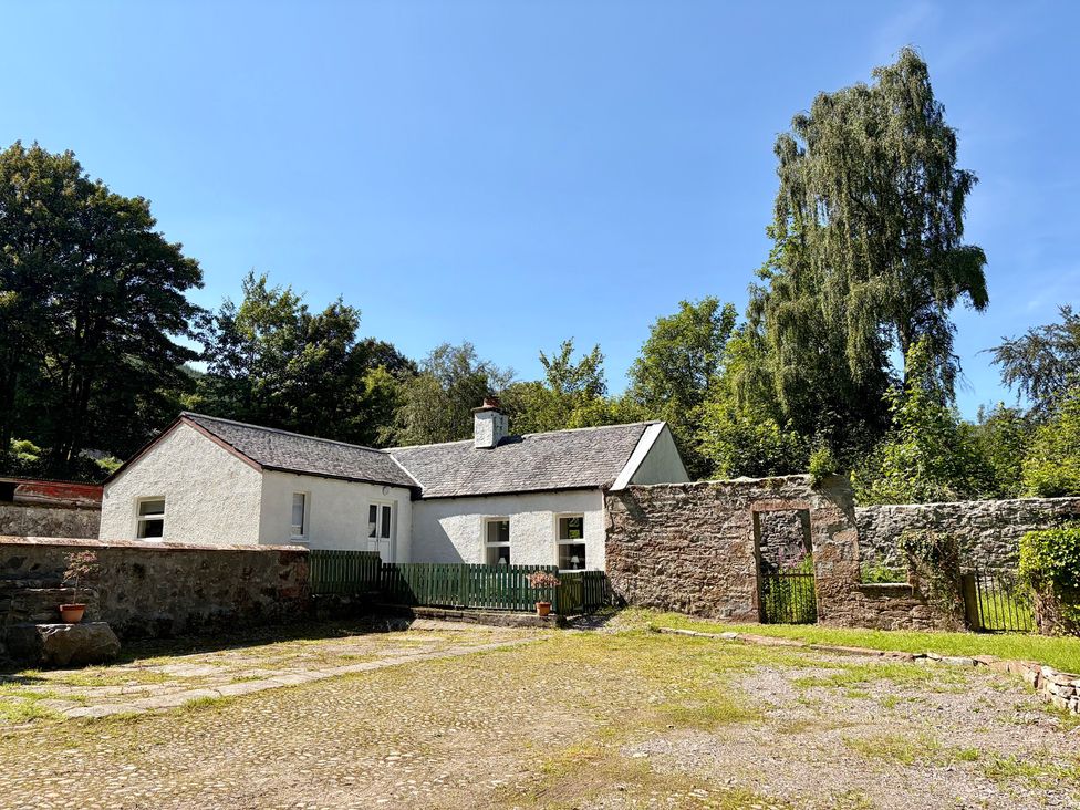 A cottage with a stone wall and trees at Laundry Cottage in Strathpeffer