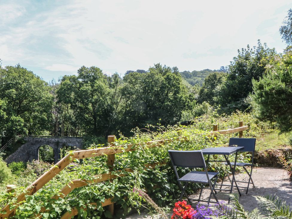 A garden with a table and chairs overlooking a bridge and trees at The Mill Barn in Lustleigh