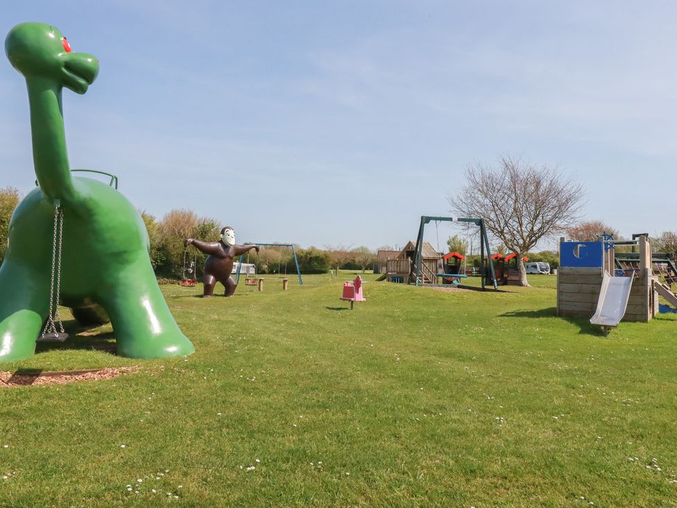 A playground with a dinosaur structure, monkey structure, swing set, slide, and playhouse at Beech Lodge in Wadebridge