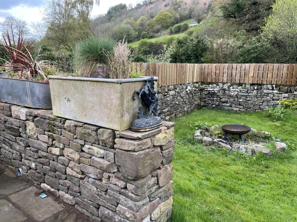 A garden with a stone wall, flower pots, a statue and a fire pit at Transformed Railway Wagon- Sleeps2-Garden-Firepit in Powys near Crickhowell