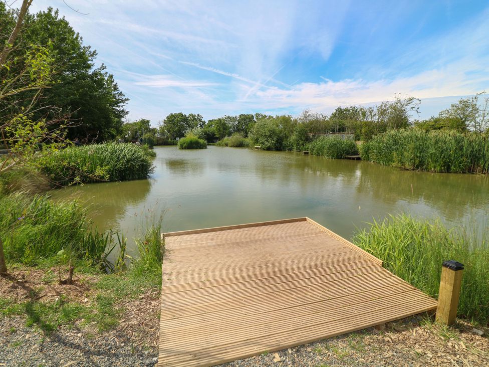 A view of a pond with a wooden deck and surrounding vegetation at Moorhen Lodge in Market Rasen