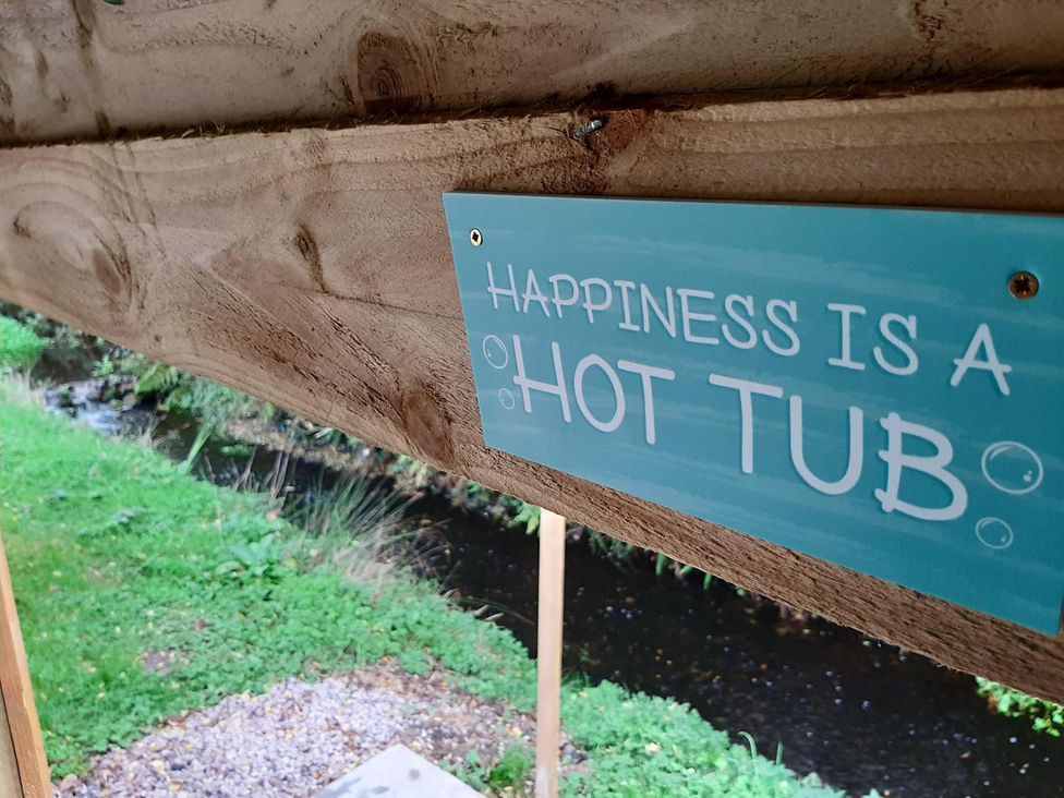 A sign that says 'Happiness is a Hot Tub' in an outdoor area near a stream and grass at Min yr Afon Cabin Sleeps2 Hot Tub Parking in Gwalchmai