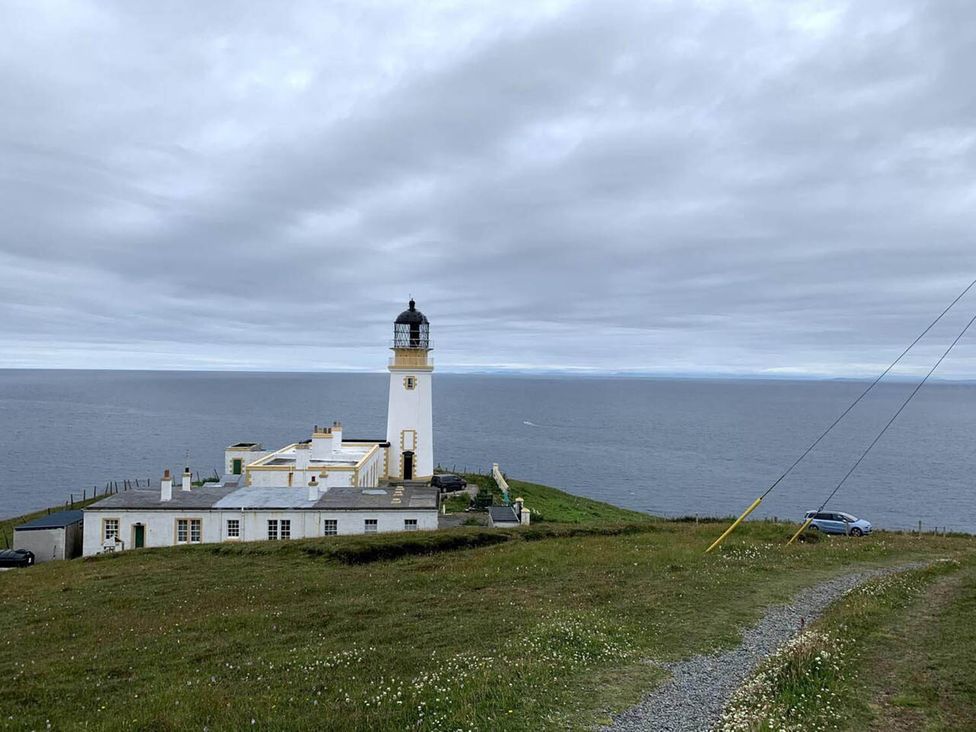 A lighthouse by the sea with a building nearby and a car parked at a coastal location