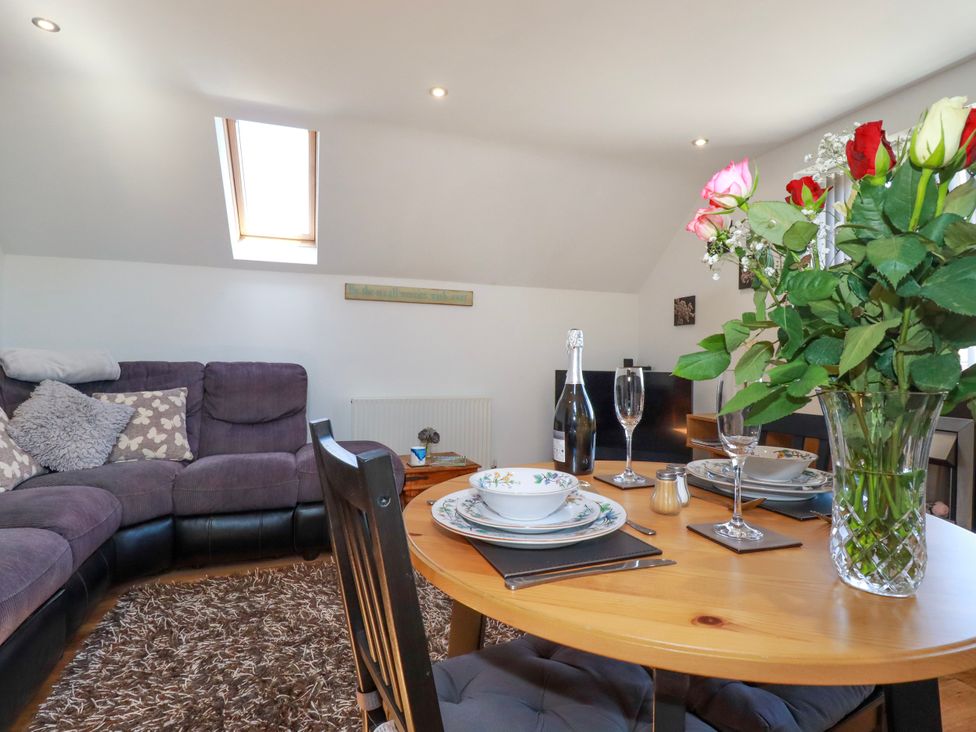 A dining area with a wooden table set with plates glasses and flowers next to a living room with a purple corner sofa and a rug at Sunnyvale in Bugle near St Austell