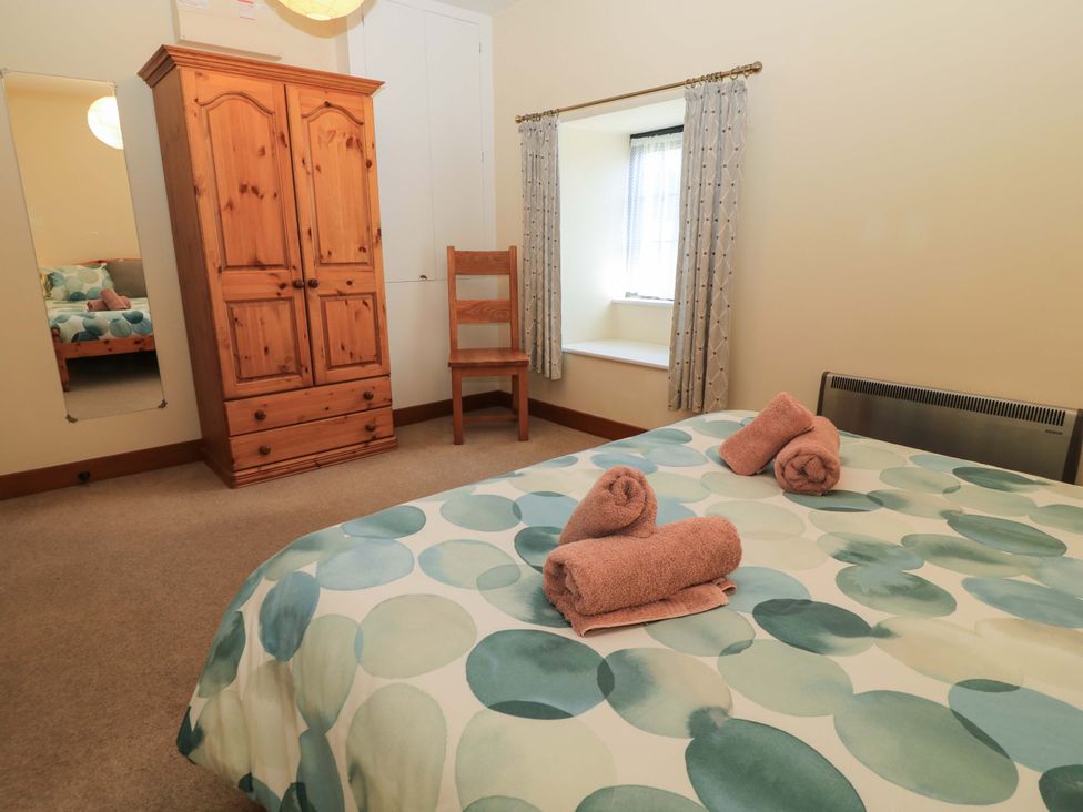 A bedroom with a bed with patterned bedding and rolled towels a wooden wardrobe a wooden chair a window with curtains and a wall mirror at Keepers Cottage in Winterborne Stickland