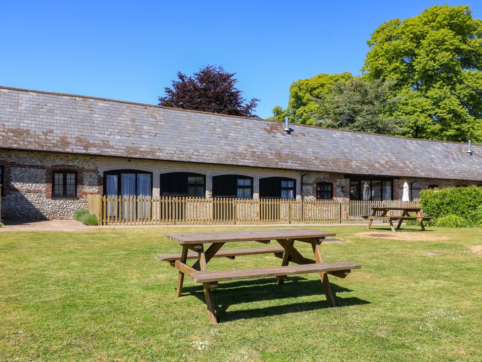 A grassy outdoor area with wooden picnic tables in front of a long stone building with black windows and a wooden fence at Keepers Cottage in Winterborne Stickland