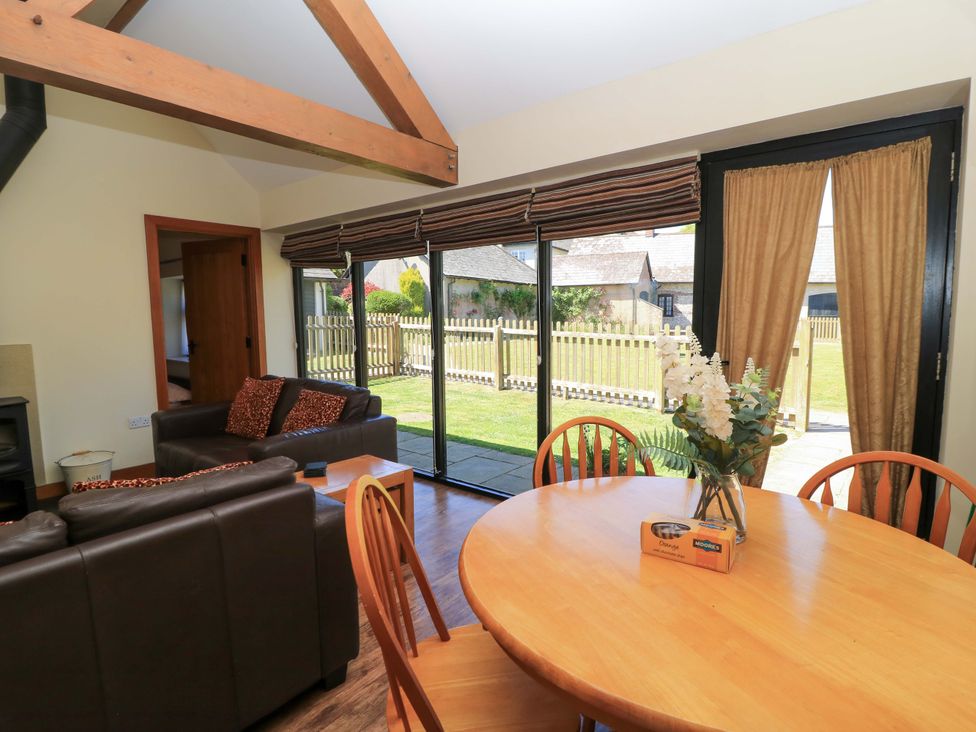 A living room with leather sofas, a wooden dining table with chairs, a vase with flowers, and large glass doors overlooking a garden at New Barn in Winterborne Stickland