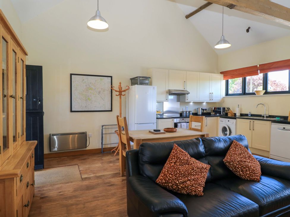 An open-plan kitchen and living area with a black sofa and patterned cushions a wooden dining table and chairs kitchen cabinets appliances and windows at The Old Cart Shed in Winterborne Stickland