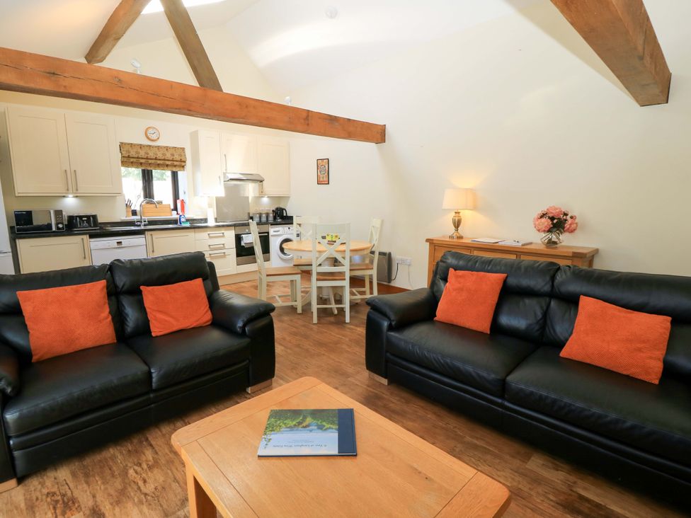 An open living area with two black leather sofas with orange cushions a wooden coffee table a round dining table with four chairs a kitchen with white cabinets and appliances and exposed wooden beams at Rowdens Barn in Winterborne Stickland