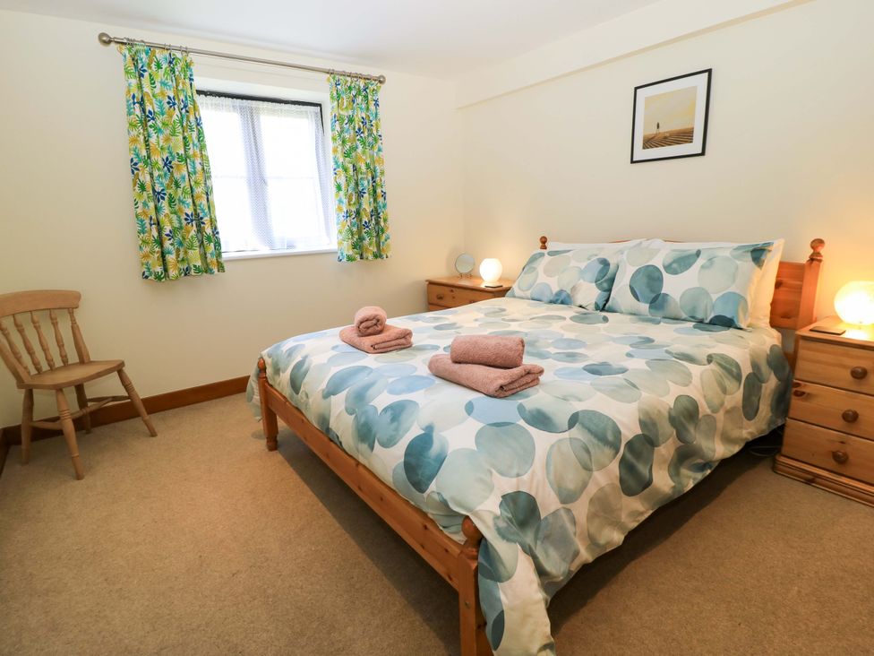 A bedroom with a bed with patterned bedding two bedside tables with lamps a wooden chair and a window with patterned curtains at Rowdens Barn in Winterborne Stickland