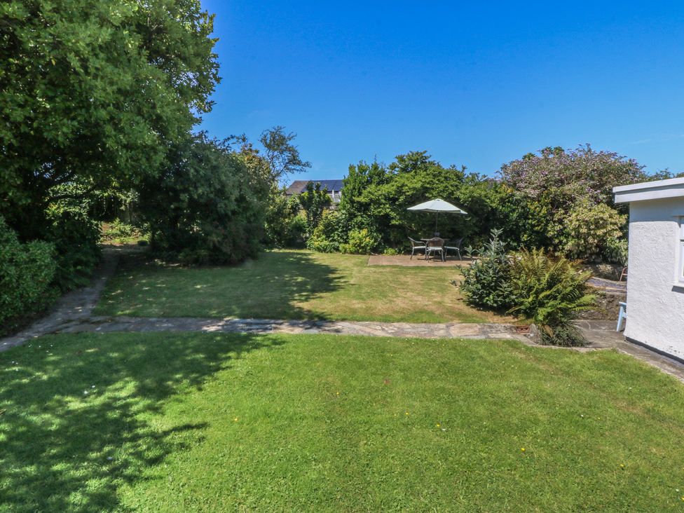A garden with grass, a pathway, trees, and a seating area with a table and umbrella at Salty Puffin in Benllech