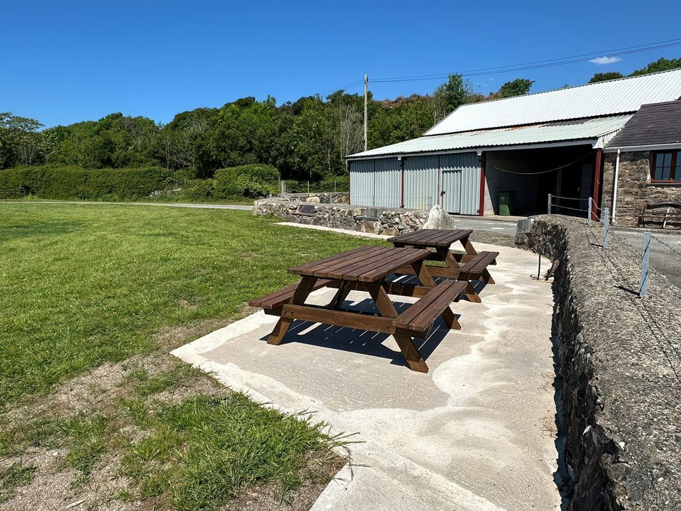 Two wooden picnic tables on a concrete patio near a stone wall and a metal building with a grassy field and trees in the background at The Mill in Abererch near Pwllheli