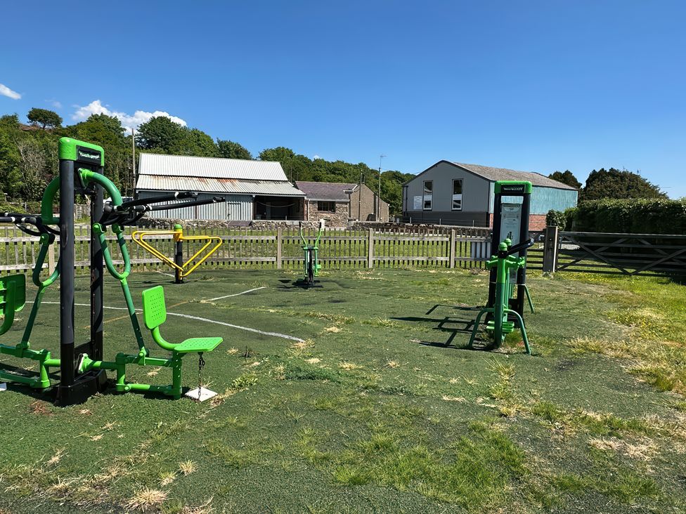 An outdoor exercise area with green fitness equipment and a fenced yard with buildings in the background at The Mill in Abererch near Pwllheli