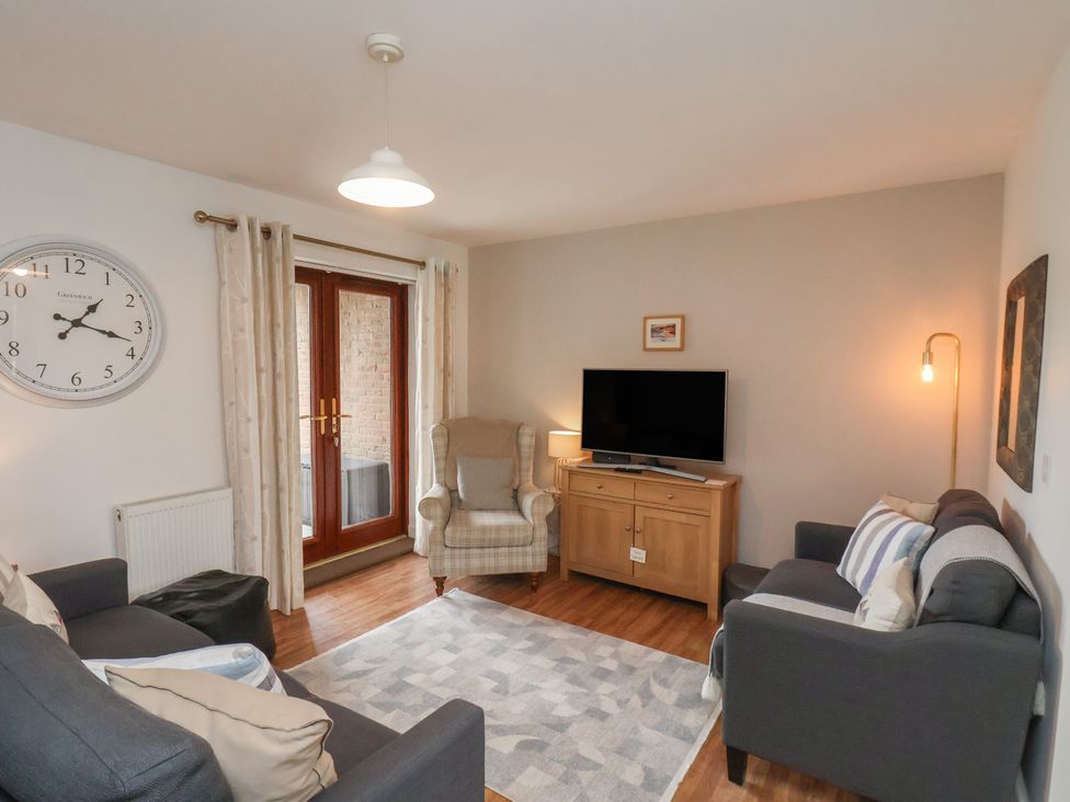 A living room with two sofas a patterned armchair a wooden cabinet with a TV a clock on the wall and curtains at Buffers in Whitby