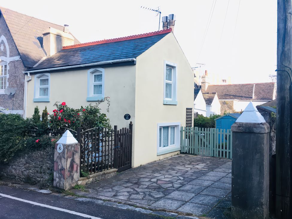 A small cream house with blue window trims and a brown gate next to a paved driveway and flowers at Little Acre in Torquay