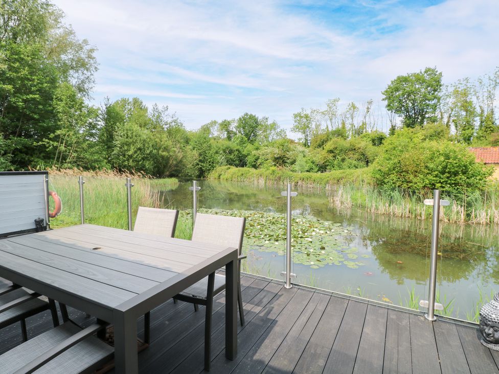 A patio with a table and chairs overlooking a pond at Dragonfly Lodge in Whinburgh near Dereham