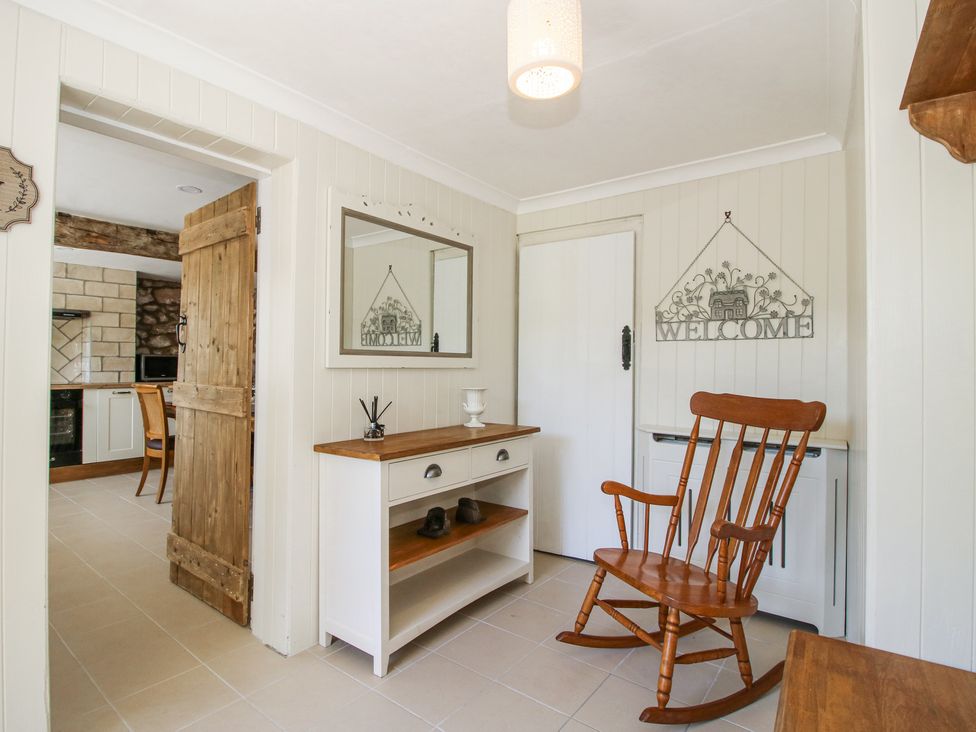 A hallway with a wooden rocking chair a white cabinet a mirror and a door with a wooden door leading to a kitchen at Pennal Cottage in Maesbury Marsh near Knockin