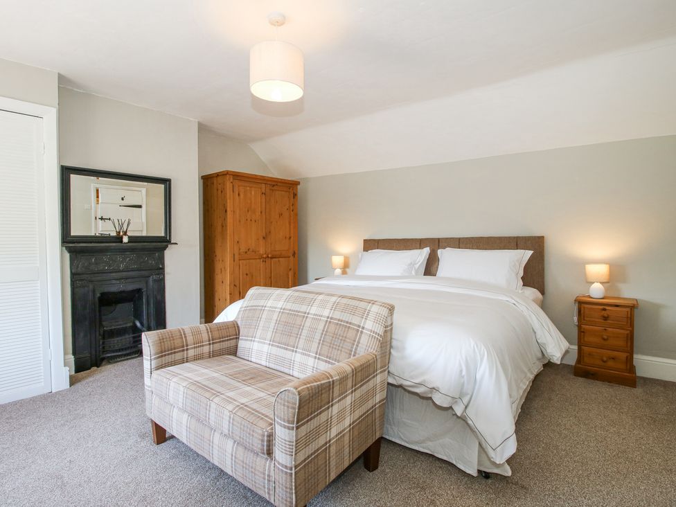 A bedroom with a double bed two bedside tables with lamps a plaid upholstered bench a wooden wardrobe and a black fireplace at Pennal Cottage in Maesbury Marsh near Knockin