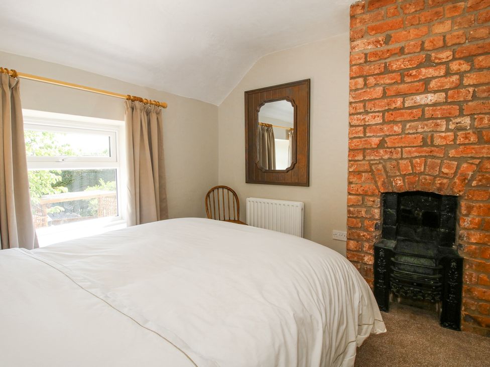 A bedroom with a bed a window beige curtains a wooden chair a mirror and a brick fireplace at Pennal Cottage in Maesbury Marsh near Knockin