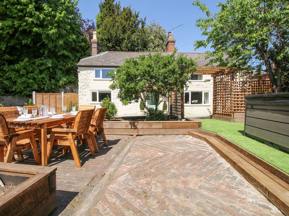 An outdoor patio area with wooden dining furniture a raised garden bed with a tree and a white house in the background at Pennal Cottage in Maesbury Marsh near Knockin