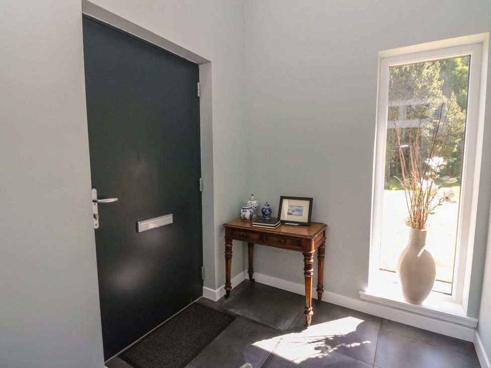 An entryway with a black door a wooden table with decorative jars books and a framed photo beside a tall window with a white vase holding dried branches at Sobrachan in Kilmartin