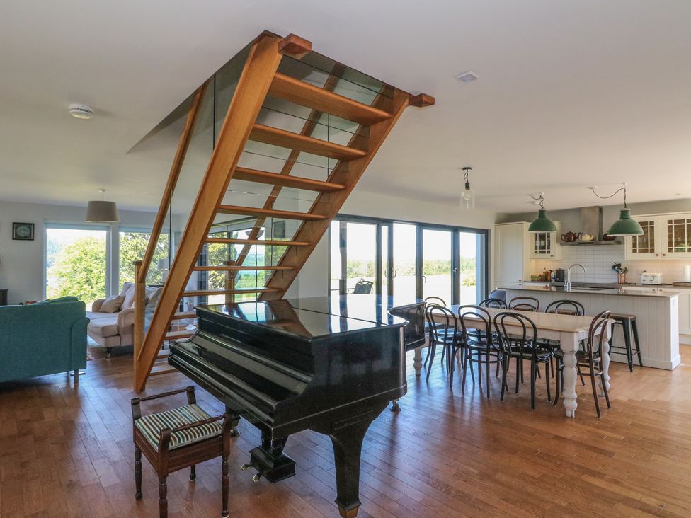 An open living space with a grand piano under wooden stairs a dining table with chairs and a kitchen area at Sobrachan in Kilmartin