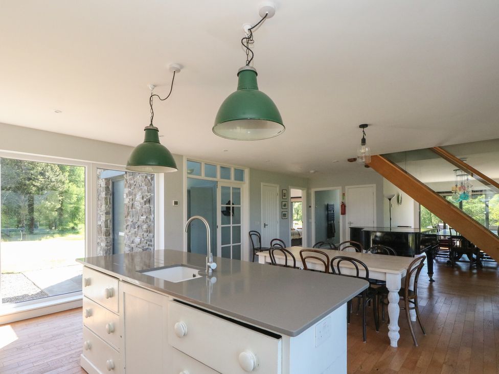 A kitchen island with a sink and green pendant lights above a dining table with chairs and a grand piano in a room with wooden floors at Sobrachan in Kilmartin