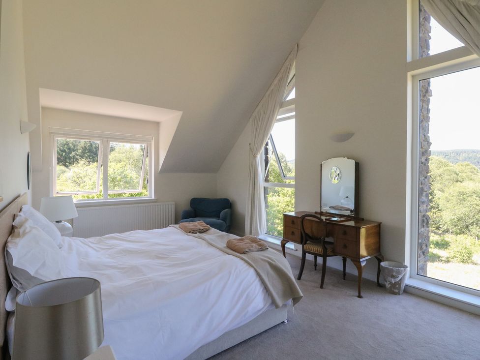 A bedroom with a bed, a chair, a dressing table with a mirror, and windows showing trees outside at Sobrachan in Kilmartin