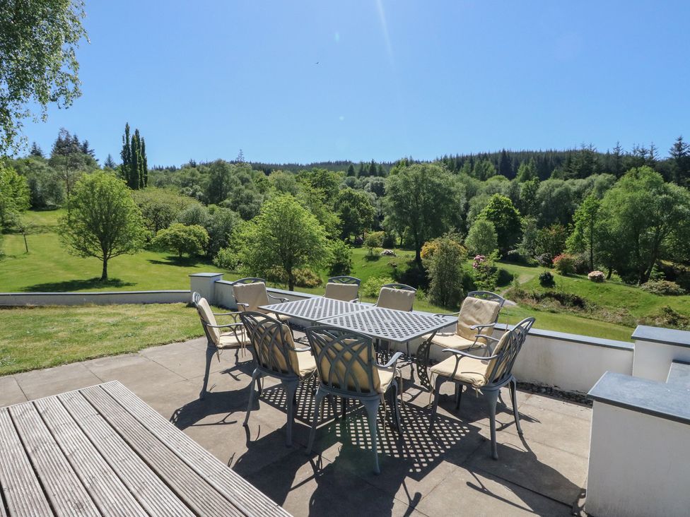 An outdoor patio with a metal dining table and eight cushioned chairs overlooking a grassy garden with trees at Sobrachan in Kilmartin