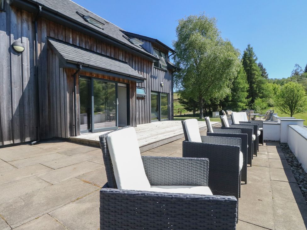 A row of cushioned outdoor chairs on a stone patio next to a wooden building with sliding glass doors and trees in the background at Sobrachan in Kilmartin