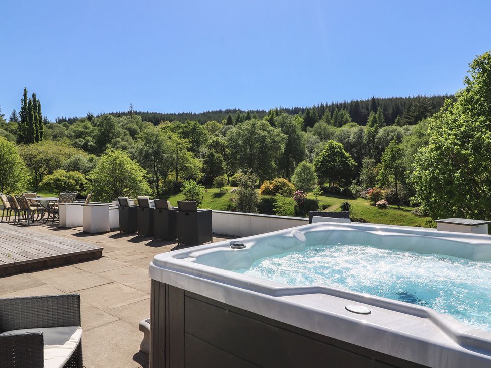 An outdoor patio area with a hot tub, chairs, and a table overlooking green trees and hills at Sobrachan in Kilmartin
