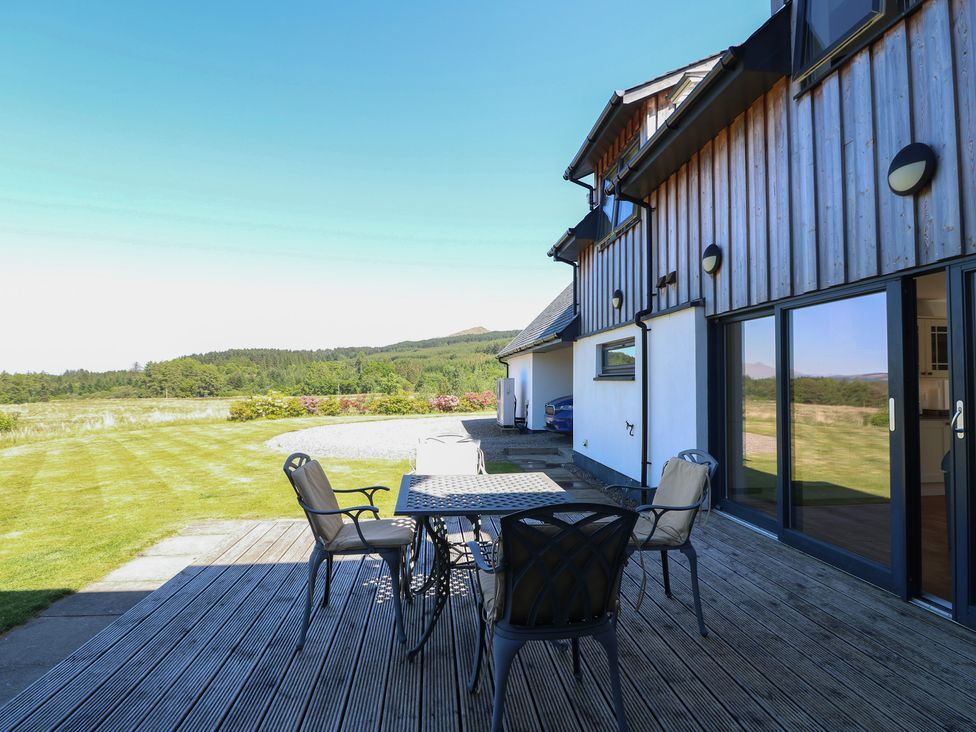 An outdoor patio with a metal table and four chairs on a wooden deck beside a modern house with sliding glass doors and a lawn with trees and hills in the background at Sobrachan in Kilmartin