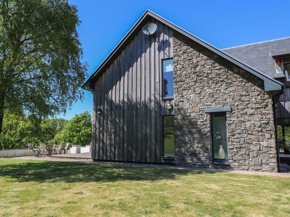 A stone and wood exterior wall of a house with windows and a satellite dish with a lawn and outdoor seating area at Sobrachan in Kilmartin