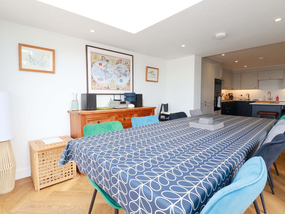 A dining room with a long table covered by a patterned tablecloth surrounded by chairs and a wooden sideboard with framed maps on the wall at Pink House in Mevagissey